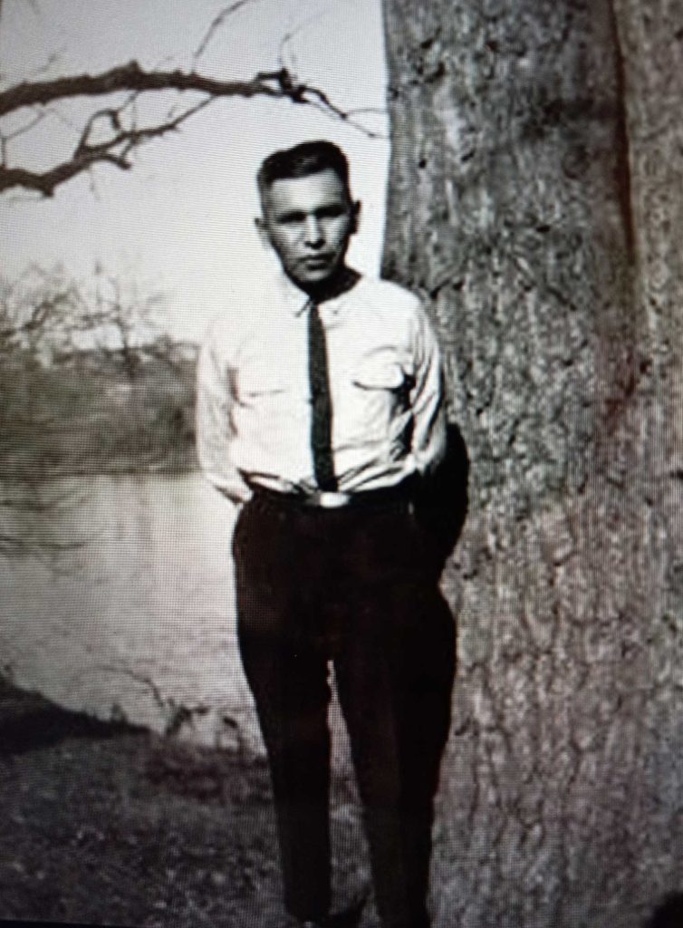 Photo of John A. Bracklin, standing by a tree, around 1930. It depicts a young man in a shirt and tie standing in front of a tree and lake. Photo courtesy of Sylvia Bracklin-Baker.